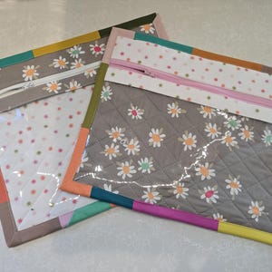 May include: Two quilted pouches with clear vinyl fronts. One pouch has a gray background with white daisies, the other has a white background with small red and green stars. Both have colorful borders and zippers.