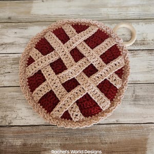 May include: A round, crocheted potholder designed to look like a cherry pie with a deep red center and a beige lattice crust. The circular piece features a beige border and a small, light-colored wooden ring attached to the side for hanging.