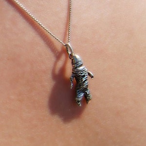May include: A silver necklace with a small, detailed astronaut pendant. The astronaut is silver and has intricate detailing. The chain is delicate and hangs against a person's skin.