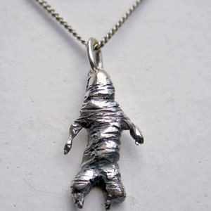 May include: A silver-toned pendant necklace featuring a stylized mummy figure. The pendant has a textured, wrapped appearance with outstretched arms and legs. The chain is a delicate, silver-colored curb chain.