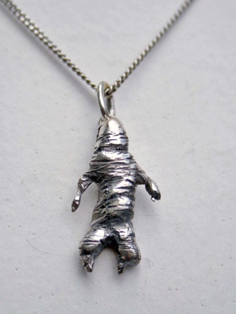 May include: A silver-toned pendant necklace featuring a stylized mummy figure. The pendant has a textured, wrapped appearance with outstretched arms and legs. The chain is a delicate, silver-colored curb chain.