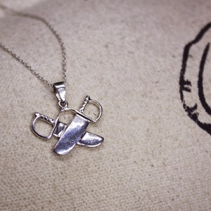 May include: A silver pendant necklace featuring a crossed swords design. The pendant has two detailed swords with curved handles and blades, suspended from a delicate chain. The necklace is displayed on a neutral-toned fabric.