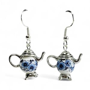 May include: A pair of silver-toned teapot earrings. Each earring features a small, white ceramic teapot with blue floral designs. The teapots have silver lids, spouts, and handles, and are suspended from silver fish hook ear wires.