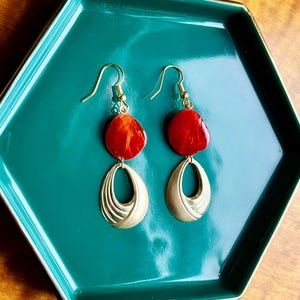 May include: A pair of dangle earrings with red, oval-shaped stones and gold-colored metal accents. The earrings are displayed on a teal hexagonal tray with a gold trim. The earrings have a hook closure.