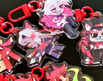 May include: A set of six colorful cartoon character keychains featuring a red, white, and blue character, a pink and white character, a red and black character, a red and white character, a red and green character, and a red and black character. The keychains are made of clear acrylic and have metal rings for attaching to keys or bags.