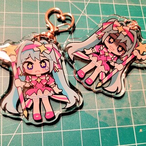 May include: Two clear acrylic keychains featuring a cartoon character with long blue hair, a pink dress, and a star-shaped wand. The keychains have a gold-colored clasp and are on a green surface.