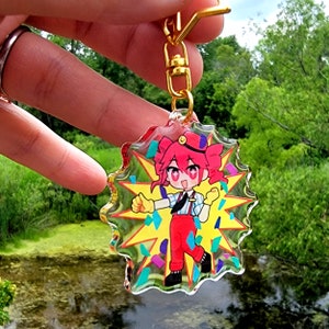 May include: A clear acrylic keychain featuring a cartoon character with red hair, a red and white outfit, and a yellow starburst background. The character is smiling and holding up both hands with a thumbs up gesture.