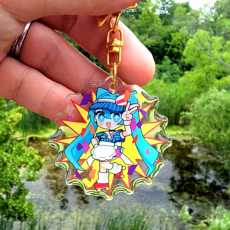 May include: A colorful anime character keychain with a blue and white hat, a blue and white dress, and blue hair. The character is holding up two fingers in a peace sign. The keychain is surrounded by confetti and a yellow starburst.