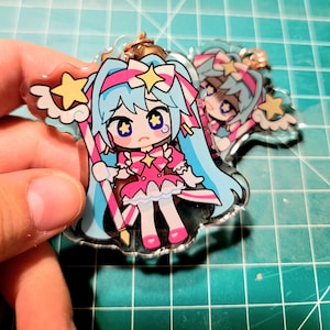May include: A clear acrylic charm featuring a cartoon character with long blue hair, a pink dress, and star accents. The charm has a gold-colored keychain attachment. The character is holding a star-topped wand and has a sad expression.