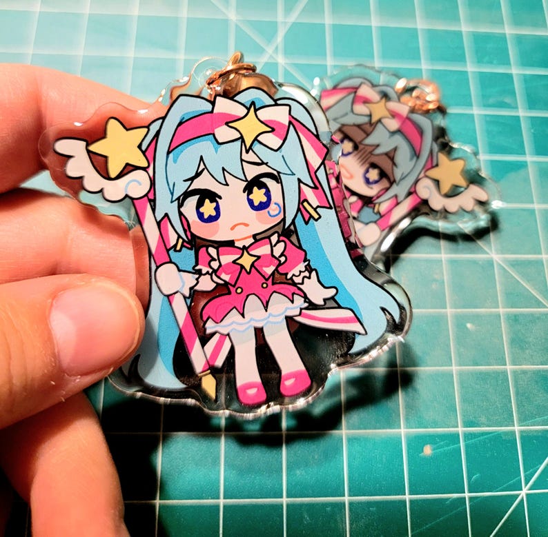 May include: A clear acrylic charm featuring a cartoon character with long blue hair, a pink dress, and star accents. The charm has a gold-colored keychain attachment. The character is holding a star-topped wand and has a sad expression.
