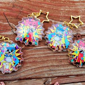 May include: Four colorful character keychains with a star-shaped charm. Each keychain features a different character with a unique design and a confetti background.