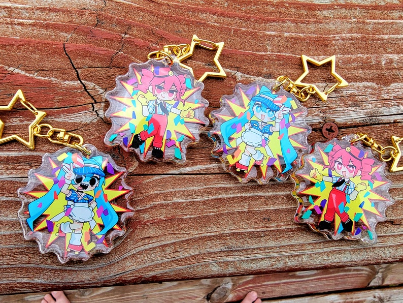 May include: Four colorful character keychains with a star-shaped charm. Each keychain features a different character with a unique design and a confetti background.