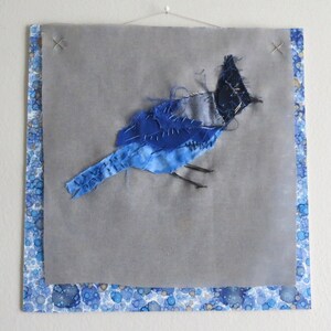 May include: A blue and gray fabric bird applique on a gray background with a blue and white patterned border.