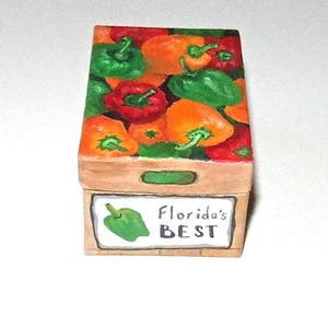 Op de afbeelding: Een houten doos met een geschilderd beeld van een doos vol groene, oranje en rode paprika's. De doos heeft een label met de tekst "Florida's BEST".