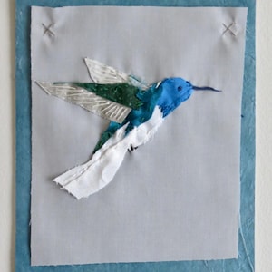 May include: A blue, green, and white fabric hummingbird is stitched onto a light gray background. The hummingbird is framed with a blue border.