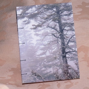 May include: A handmade journal with a misty forest scene on the cover. The cover is a light gray with a dark gray tree in the center. The tree is surrounded by other trees and fog.