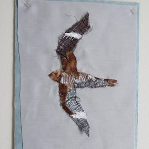 May include: A brown and white bird made of fabric is mounted on a light blue background. The bird is in flight with its wings spread out.