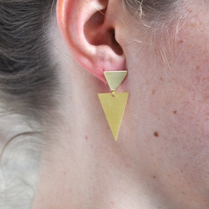 Könnte beinhalten: Ein Paar goldene Dreiecksohrringe mit einem kleineren Dreieck auf einem größeren Dreieck. Die Ohrringe hängen am Ohrläppchen.