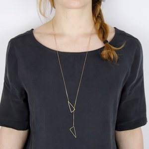 Könnte beinhalten: Eine goldene Halskette mit zwei geometrischen Dreiecksanhängern. Die Kette wird von einer Person mit langen braunen Haaren getragen.