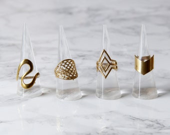 Puede incluir: Cuatro anillos de oro expuestos en soportes cónicos de acrílico transparente. Los anillos presentan diferentes diseños geométricos, incluyendo una línea ondulada, un patrón de cuadrícula, una forma de diamante y una flecha puntiaguda.