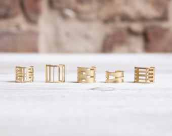 Puede incluir: Cinco anillos de color dorado con diseños geométricos. Los anillos están dispuestos en una fila sobre una superficie blanca.