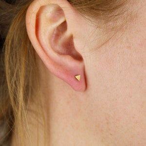 Könnte beinhalten: Nahaufnahme eines Ohrs mit einem kleinen goldenen Dreiecks-Ohrring.
