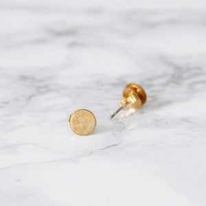 Può includere: Un paio di orecchini a bottone dorati con un design circolare. Gli orecchini sono su una superficie di marmo bianco.