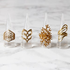 Pode incluir: Quatro anéis de ouro com diferentes desenhos geométricos, exibidos em suportes de plástico transparente. Os anéis são todos feitos de um metal dourado brilhante e têm padrões intrincados.