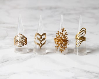 Puede incluir: Cuatro anillos de oro con diferentes diseños geométricos, expuestos en soportes de plástico transparente. Los anillos están hechos de un metal dorado brillante y tienen patrones intrincados.
