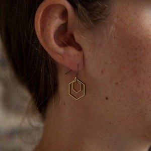 Könnte beinhalten: Goldene geometrische Ohrringe mit einem doppelten Hexagon-Design. Die Ohrringe hängen an einem Ohr einer Person.