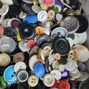 May include: A collection of various buttons in different sizes, shapes, and colors. The buttons include black, white, blue, red, and tan. Some buttons have two holes, while others have four. The buttons are made of plastic and other materials.