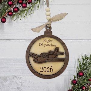 May include: Wooden Christmas ornament with a circular design. The ornament features a light-colored airplane cutout within a dark brown wooden frame. Text on the ornament reads "Flight Dispatcher" and "2026". The ornament is suspended by a twine and wooden bead hanger.