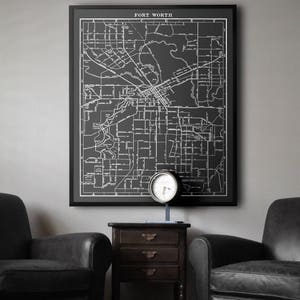 Op de afbeelding: Een zwart-witte kaart van Fort Worth, Texas, ingelijst in een zwart frame, hangt aan een grijze muur. Twee zwarte leren fauteuils staan ​​aan weerszijden van een houten bijzettafel met een klok erop.