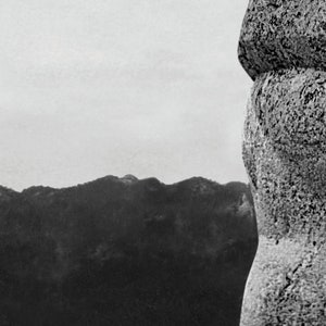 May include: A close-up of a large, grey rock formation with a rough, textured surface. The rock appears to be part of a larger structure, possibly a mountain or a statue. The background is a blurred, grey sky.