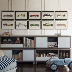 K&ouml;nnte beinhalten: Ein wei&szlig;es B&uuml;cherregal mit einer grauen Oberseite und Regalen, die mit B&uuml;chern gef&uuml;llt sind. &Uuml;ber dem B&uuml;cherregal an der Wand befinden sich neun gerahmte Drucke von Oldtimern. Die Drucke sind in schwarzen Rahmen und zeigen Autos in verschiedenen Farben.