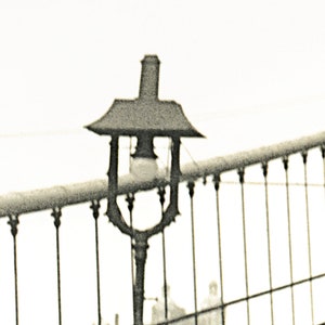 Puede incluir: Imagen en tonos sepia de una farola antigua en un puente, con una parte superior decorativa y una bombilla. La barandilla del puente y un edificio distante tambi&eacute;n son visibles, creando una est&eacute;tica cl&aacute;sica e hist&oacute;rica.