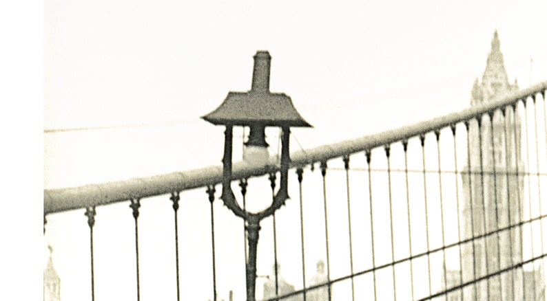 Puede incluir: Imagen en tonos sepia de una farola antigua en un puente, con una parte superior decorativa y una bombilla. La barandilla del puente y un edificio distante tambi&eacute;n son visibles, creando una est&eacute;tica cl&aacute;sica e hist&oacute;rica.