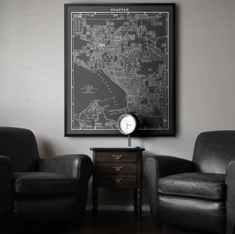 May include: A black and white vintage map of Seattle, Washington, framed in a black frame and hanging on a gray wall. Two black leather armchairs and a wooden end table with drawers are in the foreground.