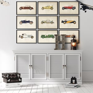 K&ouml;nnte beinhalten: Neun Vintage-Auto-Illustrationen in schwarzen Rahmen an einer wei&szlig;en Wand. Die Illustrationen sind in verschiedenen Farben gehalten und zeigen verschiedene Automodelle. Unter den Illustrationen befindet sich ein wei&szlig;er Schrank mit drei T&uuml;ren. Auf dem Schrank steht ein Buchstabe H aus Holz. Rechts neben dem Schrank steht eine Laterne.