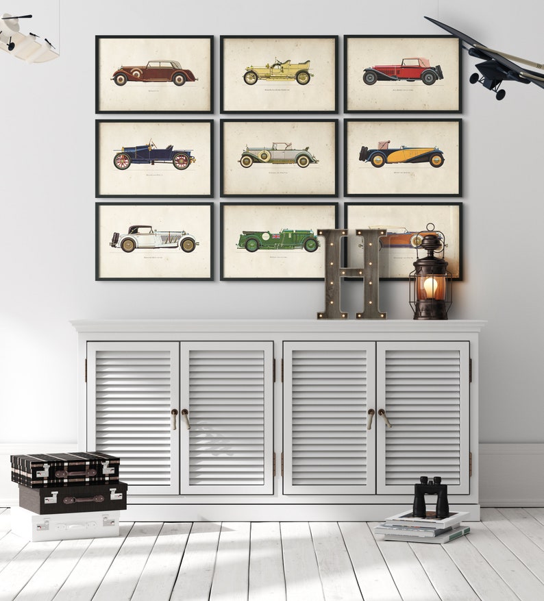 K&ouml;nnte beinhalten: Neun Vintage-Auto-Illustrationen in schwarzen Rahmen an einer wei&szlig;en Wand. Die Illustrationen sind in verschiedenen Farben gehalten und zeigen verschiedene Automodelle. Unter den Illustrationen befindet sich ein wei&szlig;er Schrank mit drei T&uuml;ren. Auf dem Schrank steht ein Buchstabe H aus Holz. Rechts neben dem Schrank steht eine Laterne.