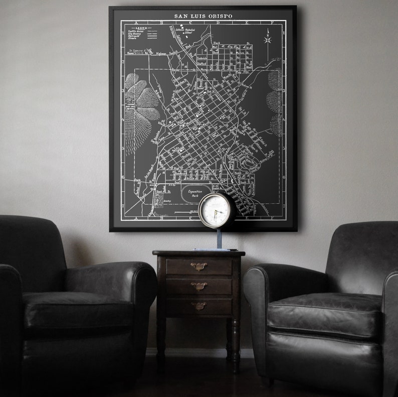Op de afbeelding: Een zwart-witte vintage kaart van San Luis Obispo, Californi&euml;, ingelijst in een zwart frame en hangend aan een grijze muur. Twee zwarte leren fauteuils staan ​​aan weerszijden van een donkerhouten bijzettafel met drie laden. Een zilveren klok staat op de tafel.