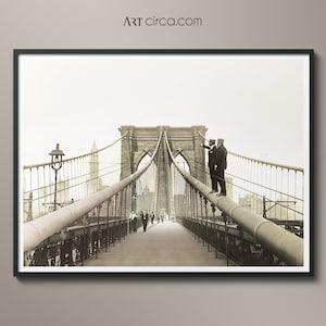 Puede incluir: Impresi&oacute;n enmarcada en tonos sepia del puente de Brooklyn. Dos hombres con trajes est&aacute;n de pie sobre una tuber&iacute;a, se&ntilde;alando. Los peatones caminan por la pasarela de madera. La imagen es de Art Circa.