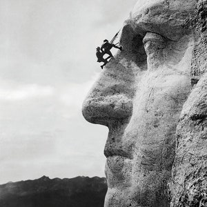 May include: A black and white photograph of Mount Rushmore National Memorial, showing the profile of George Washington's face. Two workers can be seen on the side of the mountain, working on the sculpture.