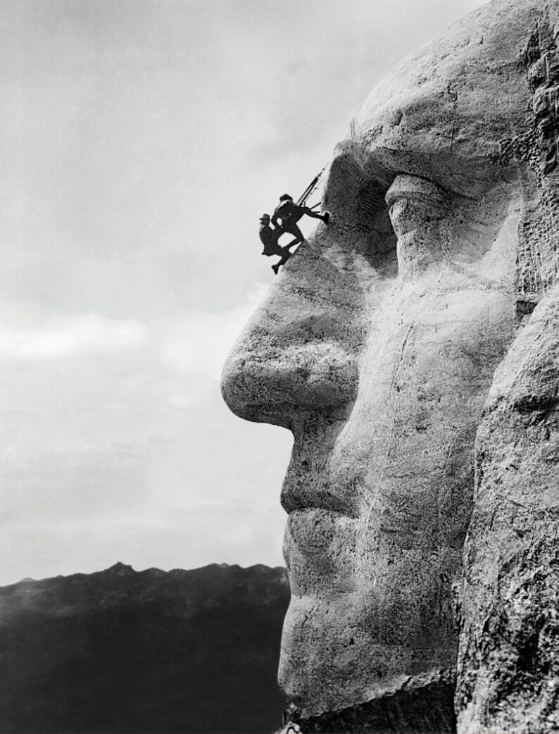 May include: A black and white photograph of Mount Rushmore National Memorial, showing the profile of George Washington's face. Two workers can be seen on the side of the mountain, working on the sculpture.