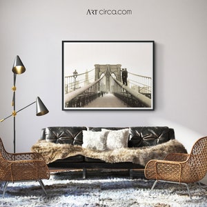 Puede incluir: Impresi&oacute;n en blanco y negro enmarcada del puente de Brooklyn sobre un sof&aacute; de cuero negro. La habitaci&oacute;n cuenta con una manta de piel sint&eacute;tica, cojines blancos, dos sillas de mimbre y una l&aacute;mpara de pie moderna. El sitio web es ARTcirca.com.