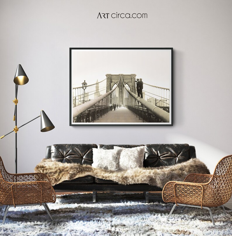 Puede incluir: Impresi&oacute;n en blanco y negro enmarcada del puente de Brooklyn sobre un sof&aacute; de cuero negro. La habitaci&oacute;n cuenta con una manta de piel sint&eacute;tica, cojines blancos, dos sillas de mimbre y una l&aacute;mpara de pie moderna. El sitio web es ARTcirca.com.