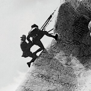 May include: Two people are climbing a large rock formation. The people are silhouetted against a bright sky. The rock formation is textured and appears to be a sculpture.