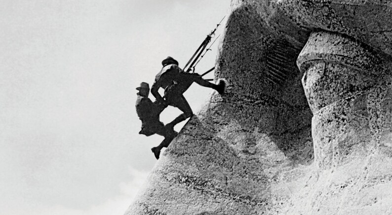 May include: Two people are climbing a large rock formation. The people are silhouetted against a bright sky. The rock formation is textured and appears to be a sculpture.