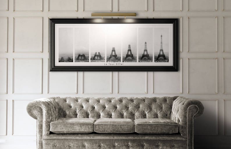 May include: Black and white framed print of the Eiffel Tower's construction phases, above a tufted gray sofa. The print is labeled "La Tour Eiffel". The wall is white with rectangular paneling, and a gold-toned light fixture is above the artwork.