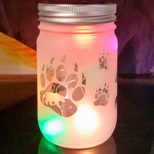 May include: A frosted glass jar with a silver lid and a solar panel on top. The jar has a bear paw design etched into the glass. The jar is lit up with colorful LED lights. The text "Bear Paw" is printed below the jar.